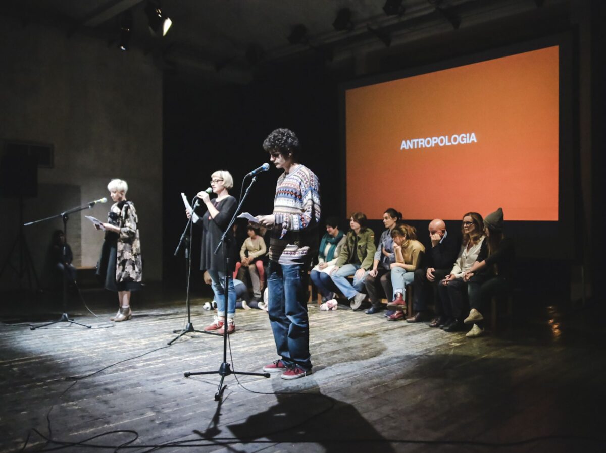 Tre persone leggono in primo piano al microfono, un gruppo di persone siede dietro di loro in ascolto, sullo sfondo proiettata sullo schermo la parola antropologia, nello spettacolo Family Affair di ZimmerFrei