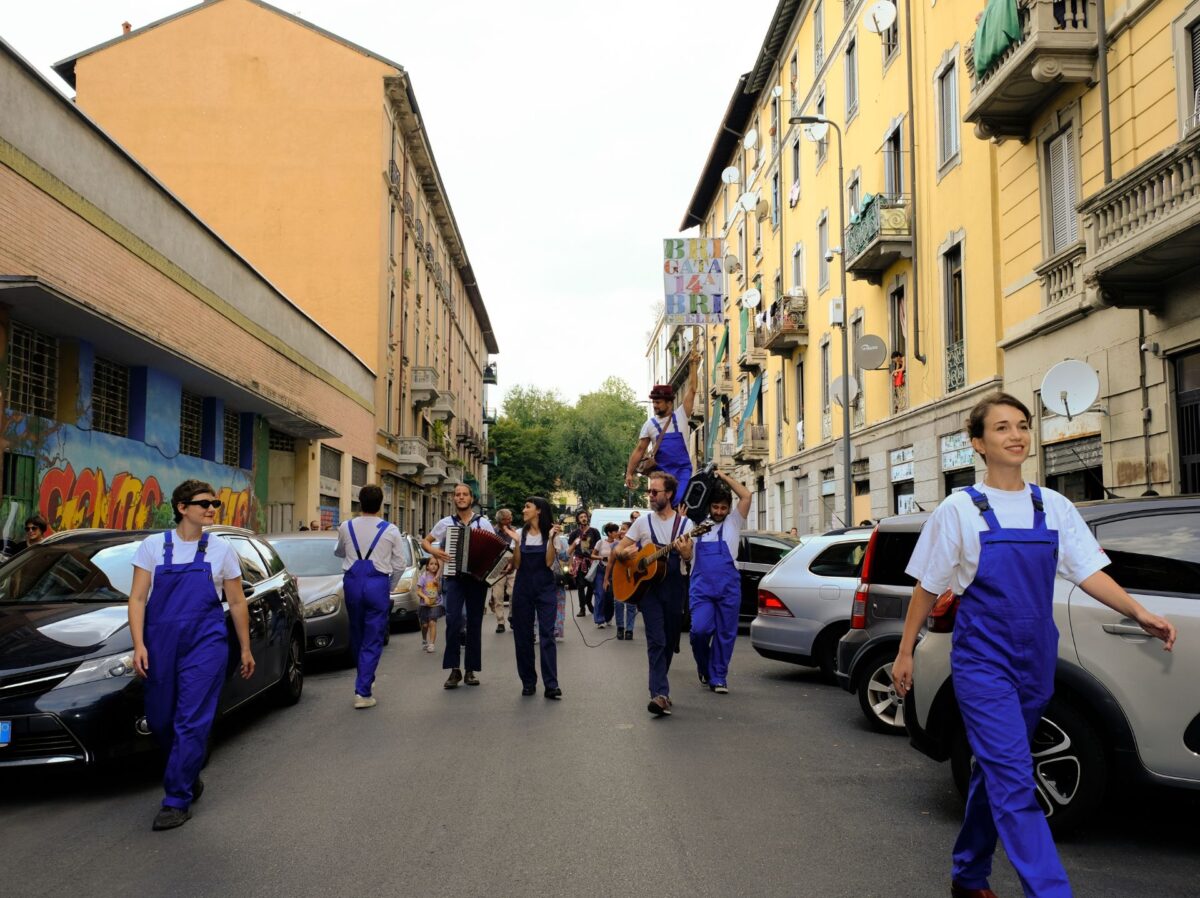 Un gruppo di musicisti, attori e trampolieri in tuta blu sfilano gioiosi nelle vie del quartiere padova/turro di milano per raccontare fiabe nei cortili dei condomini