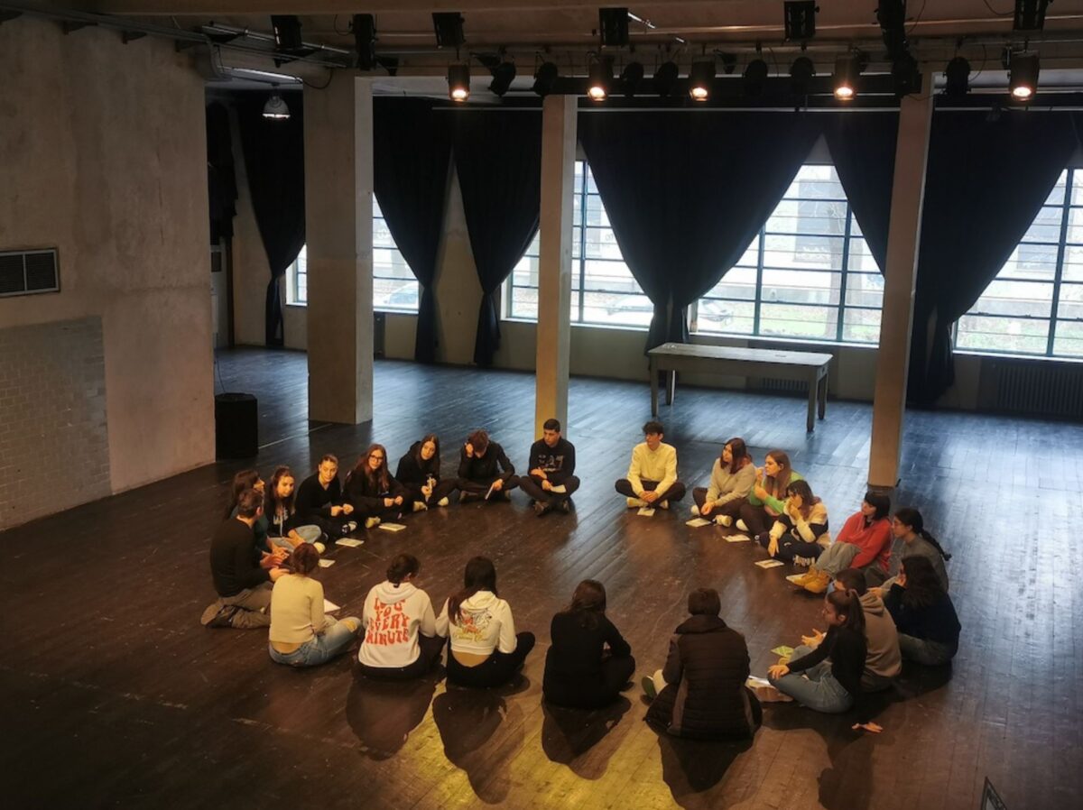 Un gruppo di studenti e studentesse siede in cerchio durante un incontro del laboratorio di scrittura e teatro il margine e il centro di ateliersi, sul fondo la vetrata del teatro con tendoni aperti