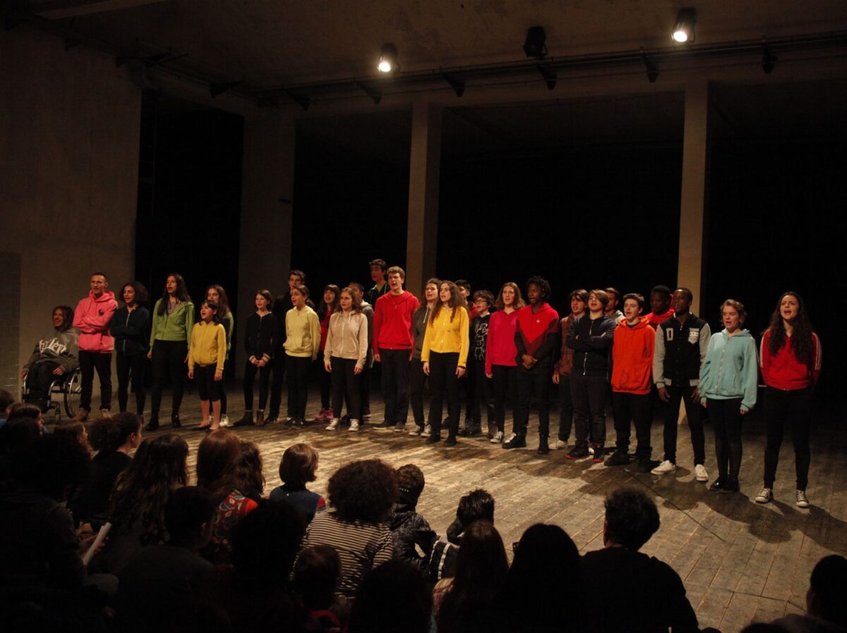 Un gruppo di ragazze e ragazzi con la magliette colorate stanno dritti di fronte al pubblico, recitando i versi, nell'esito pubblico del laboratorio di teatro non-scuola, Timone d'Atene