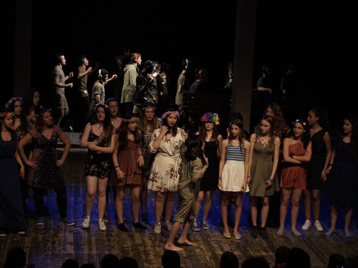 Ragazze con vestiti floreali si preparano a partecipare a una festa, sullo sfondo ragazzi in vestiti militare e con le cuffie camminano sulle panche, sono i marinai e le sirene dell'Odissea, nell'esito del laboratorio non-scuola