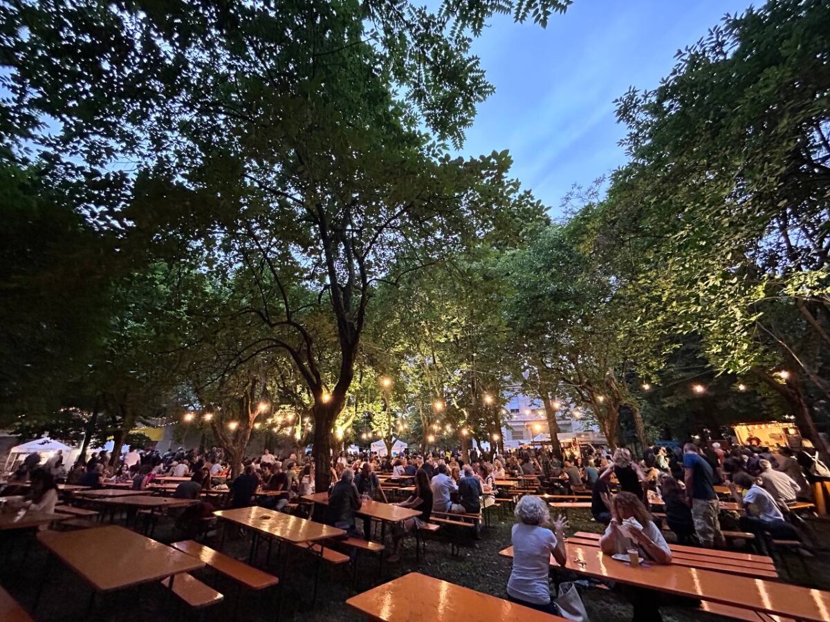 Persone mangiano e conversano al crepuscolo sotto gli alberi in un punto ristoro nel parco del Paolo Pini durante la festa di Radio Popolare