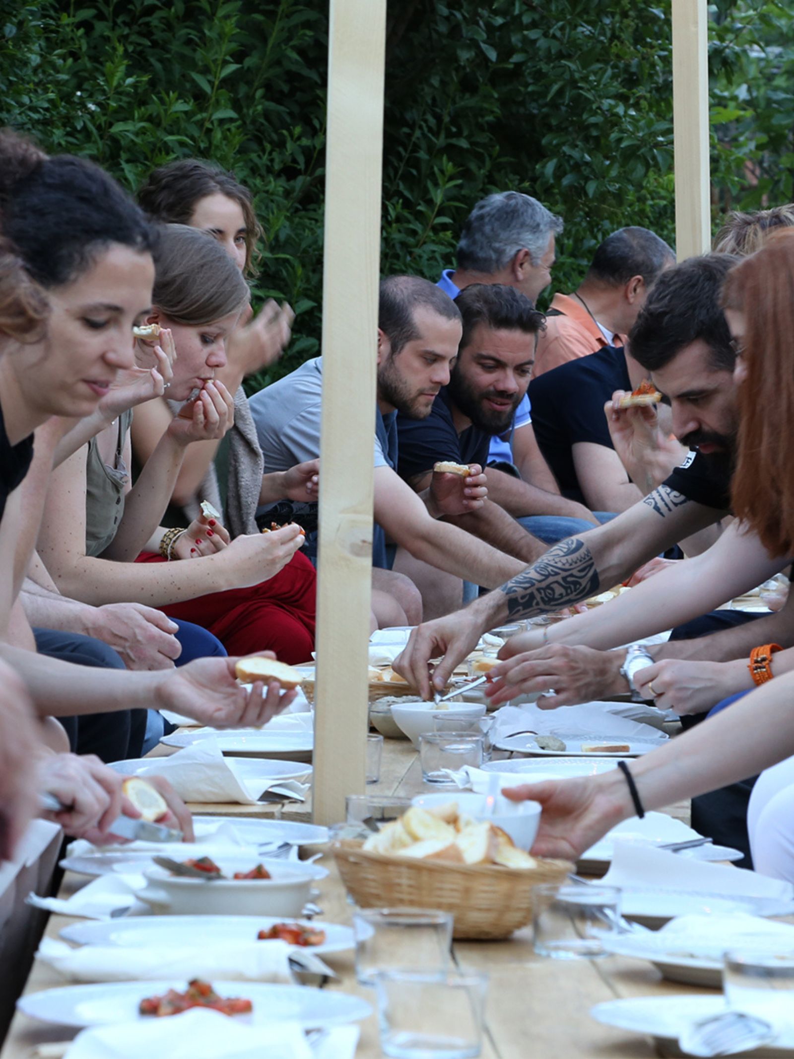 Persone condividono il cibo intorno a un tavolo, le une sedute di fronte alle altre, in una sera d'estate nel parco per il progetto speciale Un'Odissea in Comasina