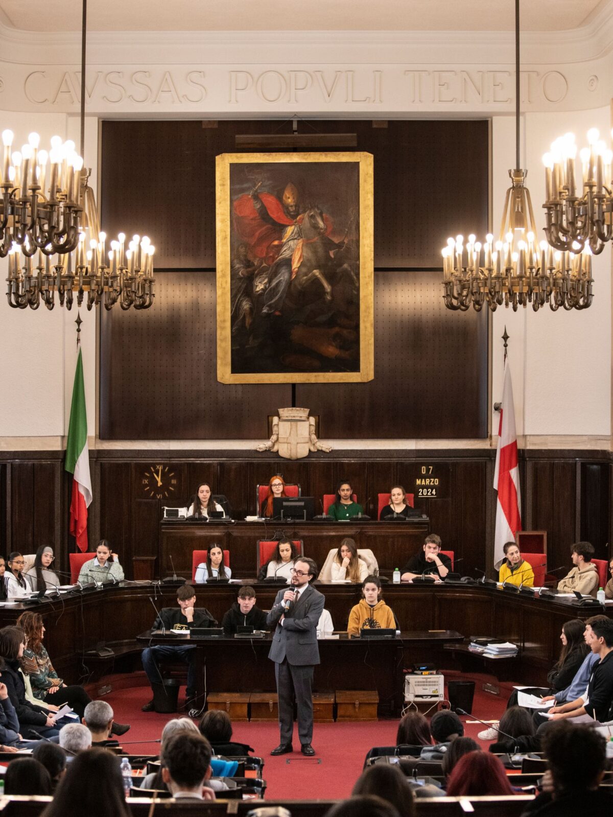 Al centro l'attore che interpreta Primo Levi, parla a studentesse e studenti seduti al posto dei consiglieri nell'aula del consiglio comunale di palazzo con marino, con maestosi lampadari e un grosso quadro sul fondo