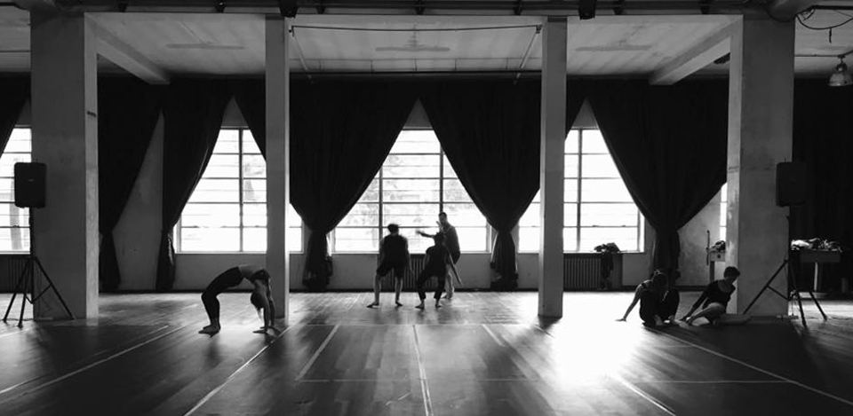 Un gruppo di danzatori prova di fronte ad un'ampia vetrata, con i tendoni aperti, in un edificio dall'architettura industriale, l'ex mensa dell'O.P. Paolo Pini, ora teatrolacucina