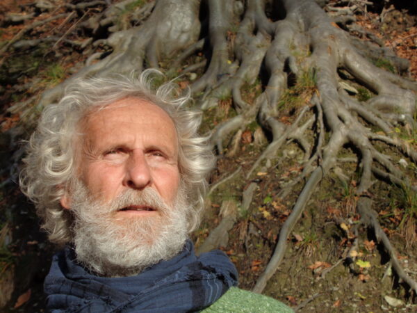 In primo piano il volto di un uomo con barba e capelli bianca, dietro le radici di un maestoso albero
