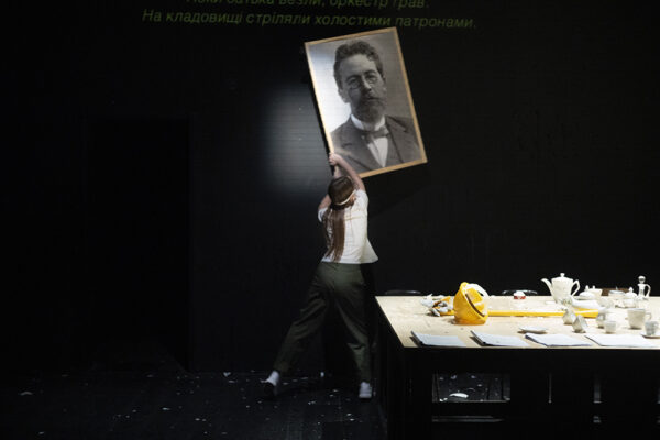 Una ragazza di schiena sta togliendo dalla parete scura un quadro che raffigura lo scrittore russo cechov, davanti a lei un tavolo con tazzine e piatti di ceramica e un elmetto giallo