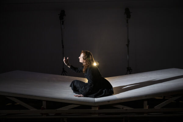 Una donna vestita di nero con i capelli rossi sciolti sta seduta su un tappeto bianco lucido, che proietta la sua ombra, lo sguardo rivolto alla mano alzata, con fare languido