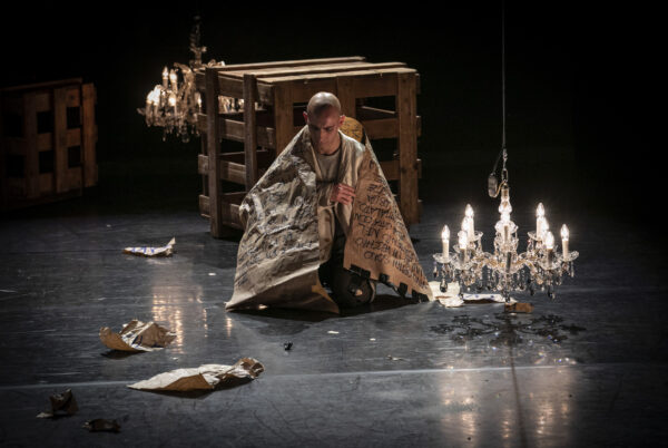 Un giovane uomo sta seduto in mezzo al palcoscenico, avvolto in una coperta, di fronte a lui un lampadario a goccia illuminato, dietro di lui una struttura in pallet di legno e intorno dei fogli di carta strappati