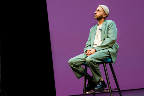 L'attore sta seduto su un trespolo, con sguardo perso, e uno sfondo violaceo