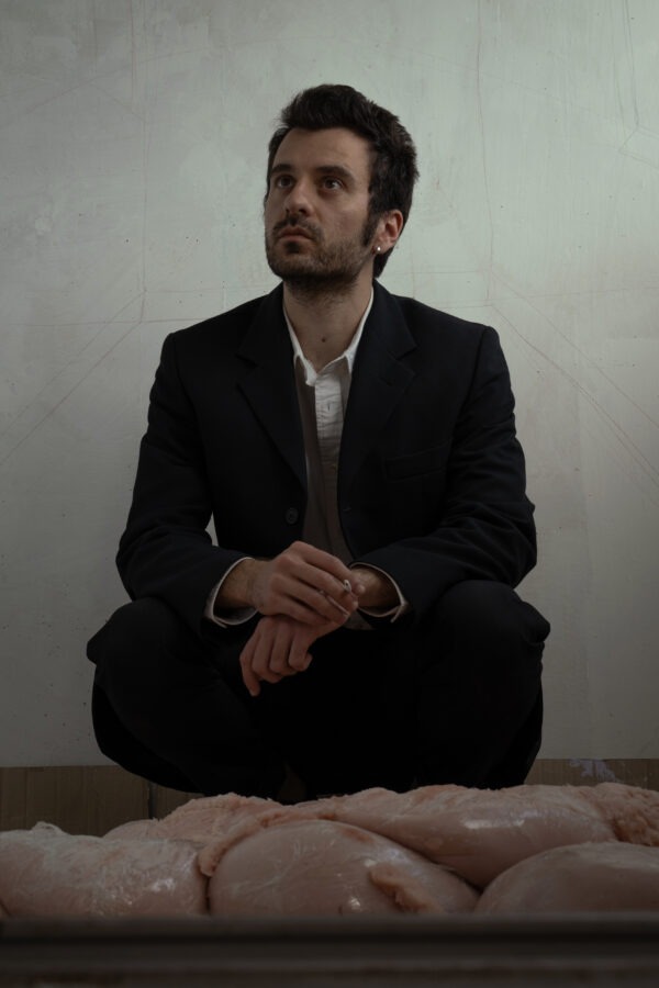 Un ragazzo accovacciato con completo nero e camicia bianca, con una sigaretta tra le mani, guarda confuso fuori dall'immagine, come se stesse accadendo qualcosa che cattura la sua attenzione, davanti a lui del materiale rosa, come fosse carne di animale.