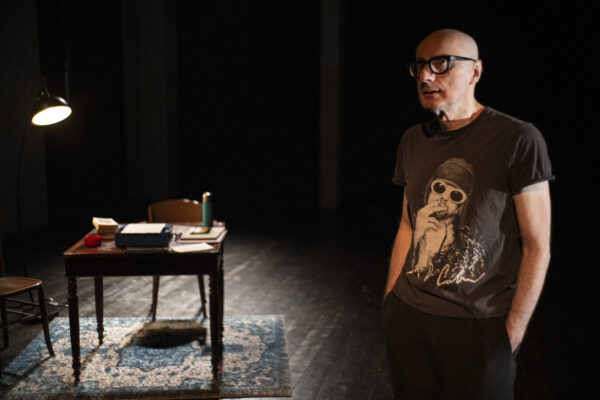 L'attore sta di fronte al pubblico, dietro una scrivania con una borraccia e dei libri, per terra un tappeto damascato,s et di uno studio letterario