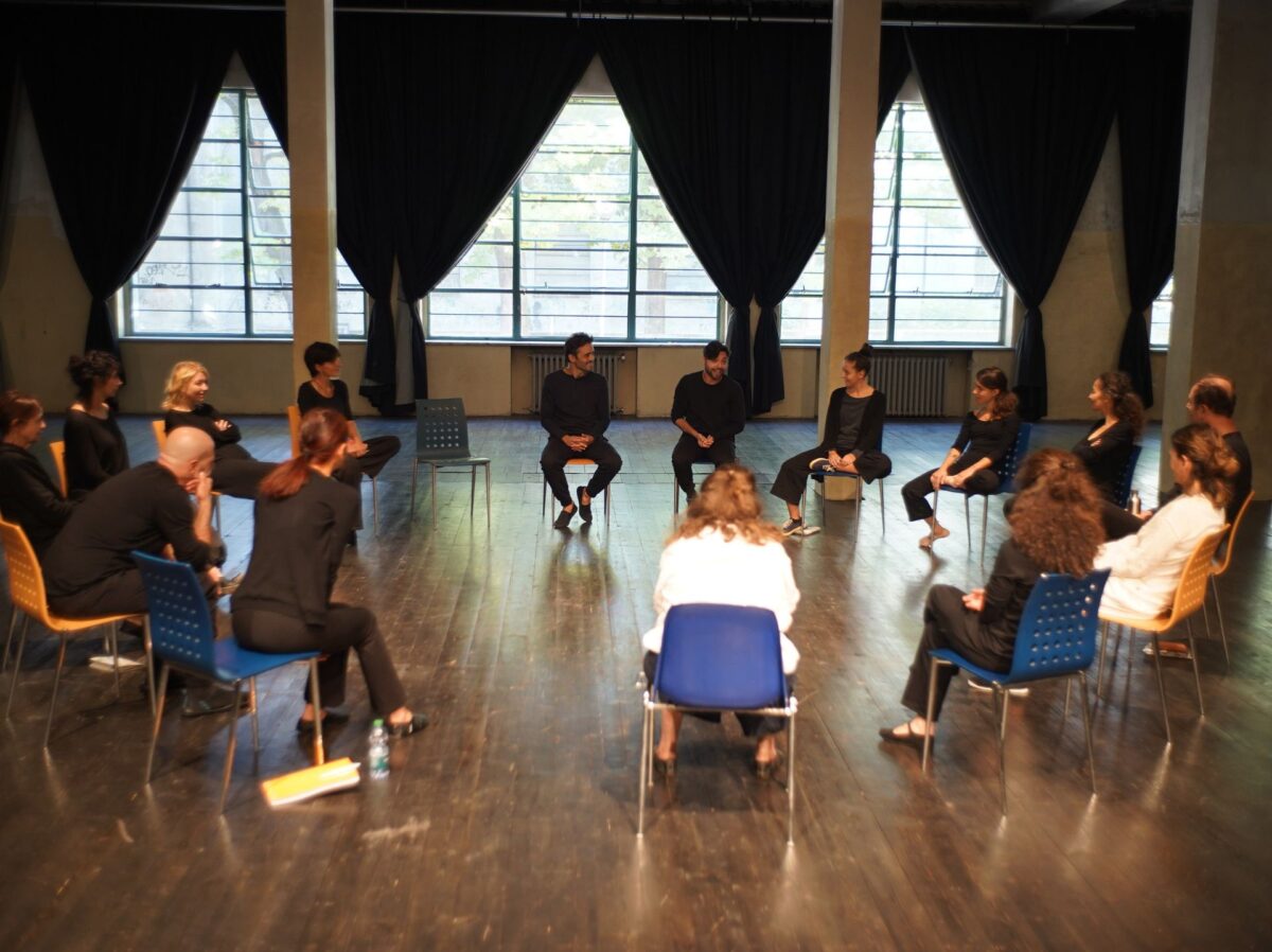 Un gruppo di attori sta seduto su sedie in mezzo alla sala del teatro, in cerchio, dietro ampie e luminose vetrate di un edificio dall'architettura industriale