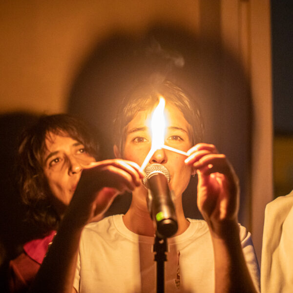 una giovane donna sta davanti a un microfono con in mano due fiammiferi accesi, dietro di lei fa capolino il volto di un'altra donna, in un'atmosfera sospesa