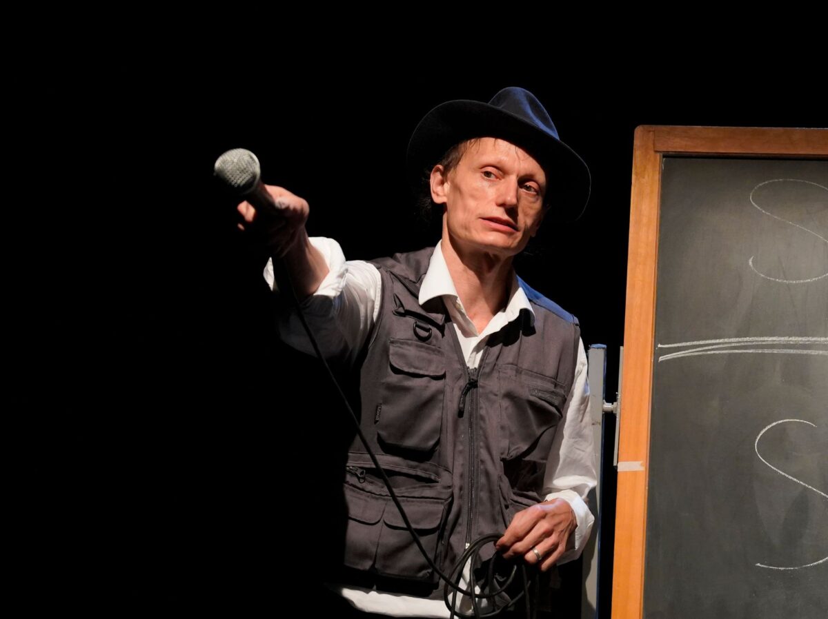 L'attore che interpreta l'artista joseph beuys rivolge un microfono verso il pubblico, con gesto performativo