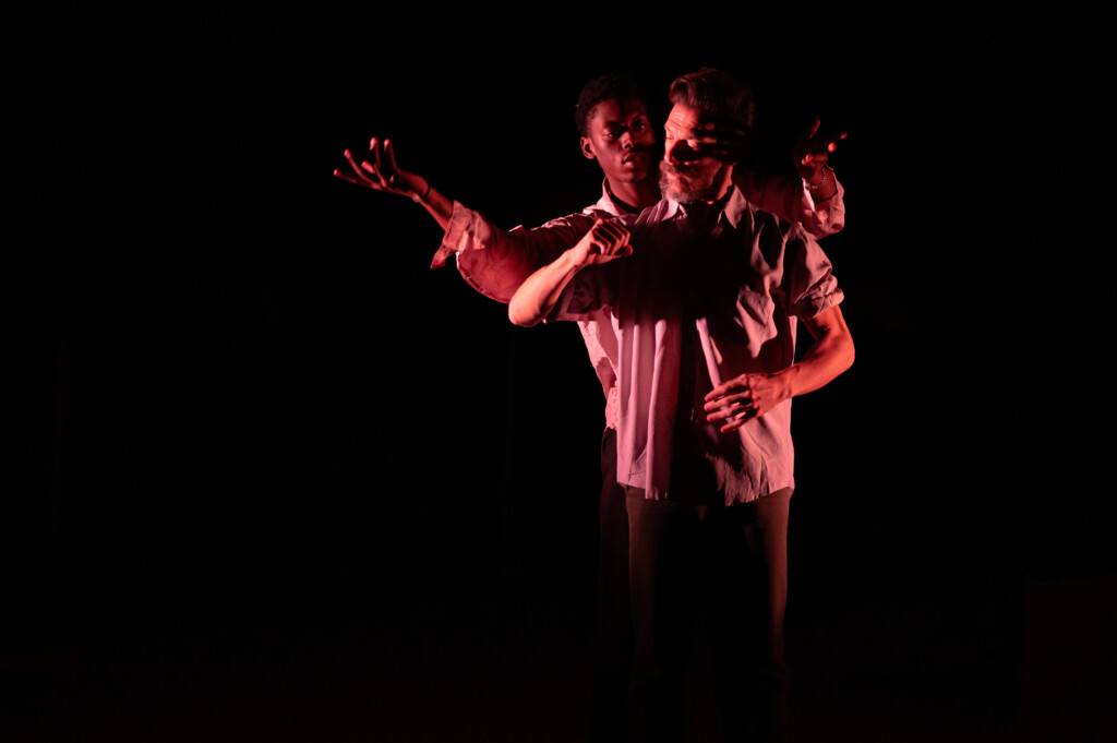 Un danzatore fa capolino poggiando la testa sull'incavo delle spalle dell'altro danzatore, in un intreccio di mani e braccia che riproduce i movimenti della coreografia a due