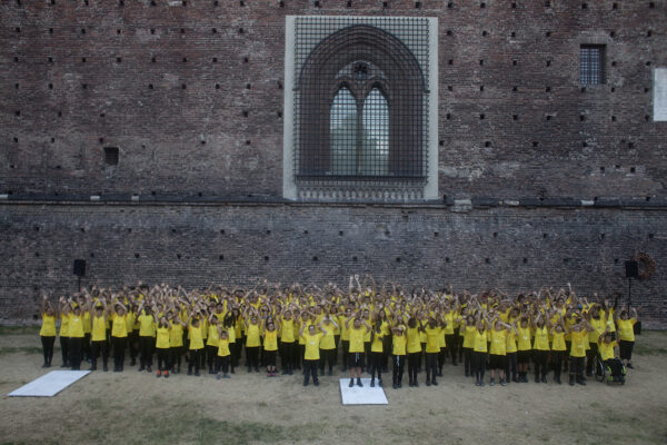 Un gruppo di ragazzi e ragazze in maglietta gialla sta compatto nel fossato del castello sforzesco, per il progetto speciale eresia della felicità