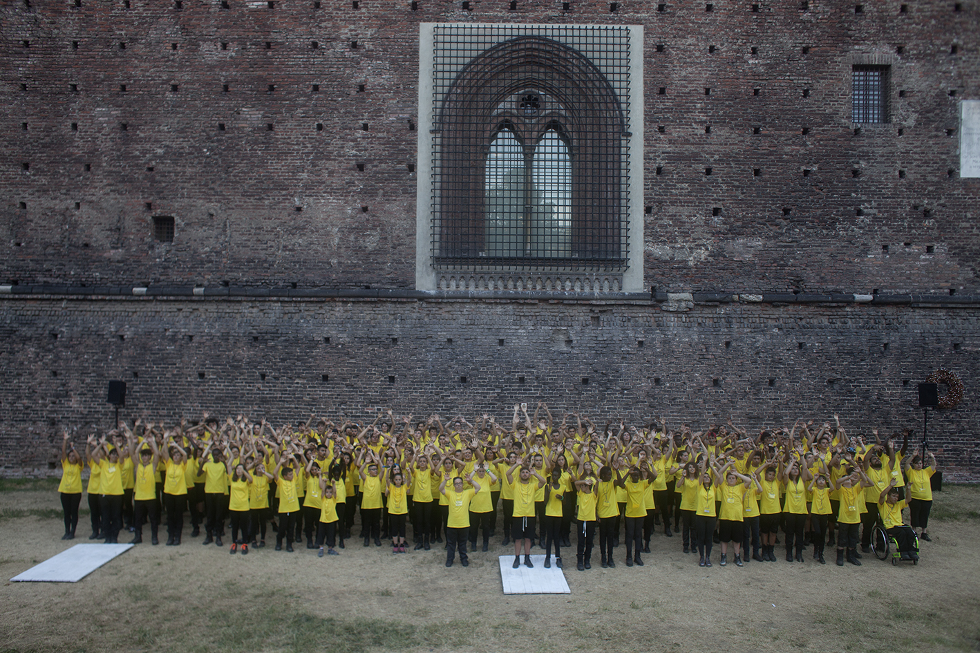 Un gruppo di ragazzi e ragazze in maglietta gialla sta compatto nel fossato del castello sforzesco, per il progetto speciale eresia della felicità