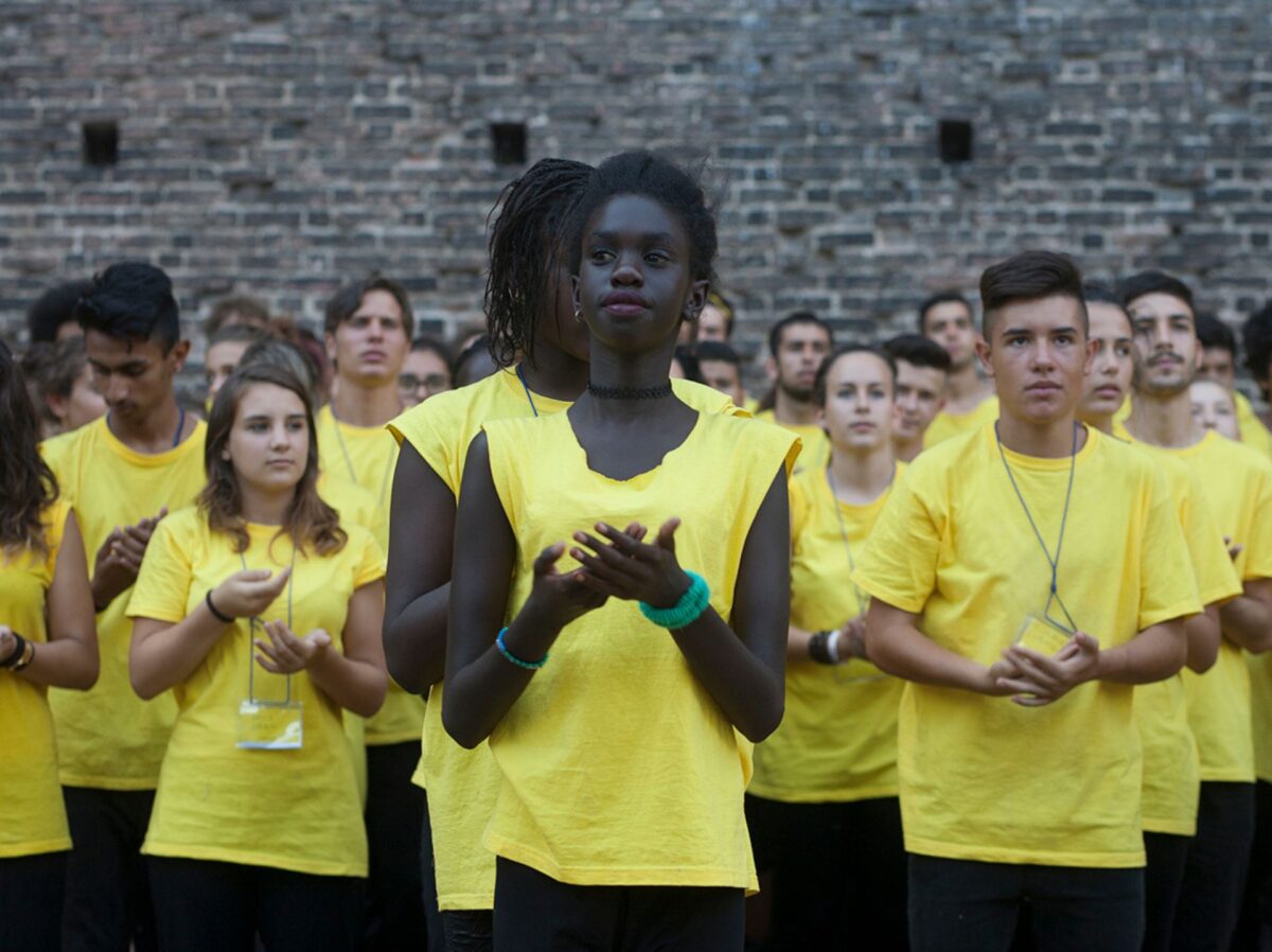 un gruppo di ragazze e ragazzi in maglietta gialla recita con concentrazione e serietà i versi del poeta dissidente russo majakovskij nel fossato del castello sforzesco di milano per il progetto speciale eresia della felicità