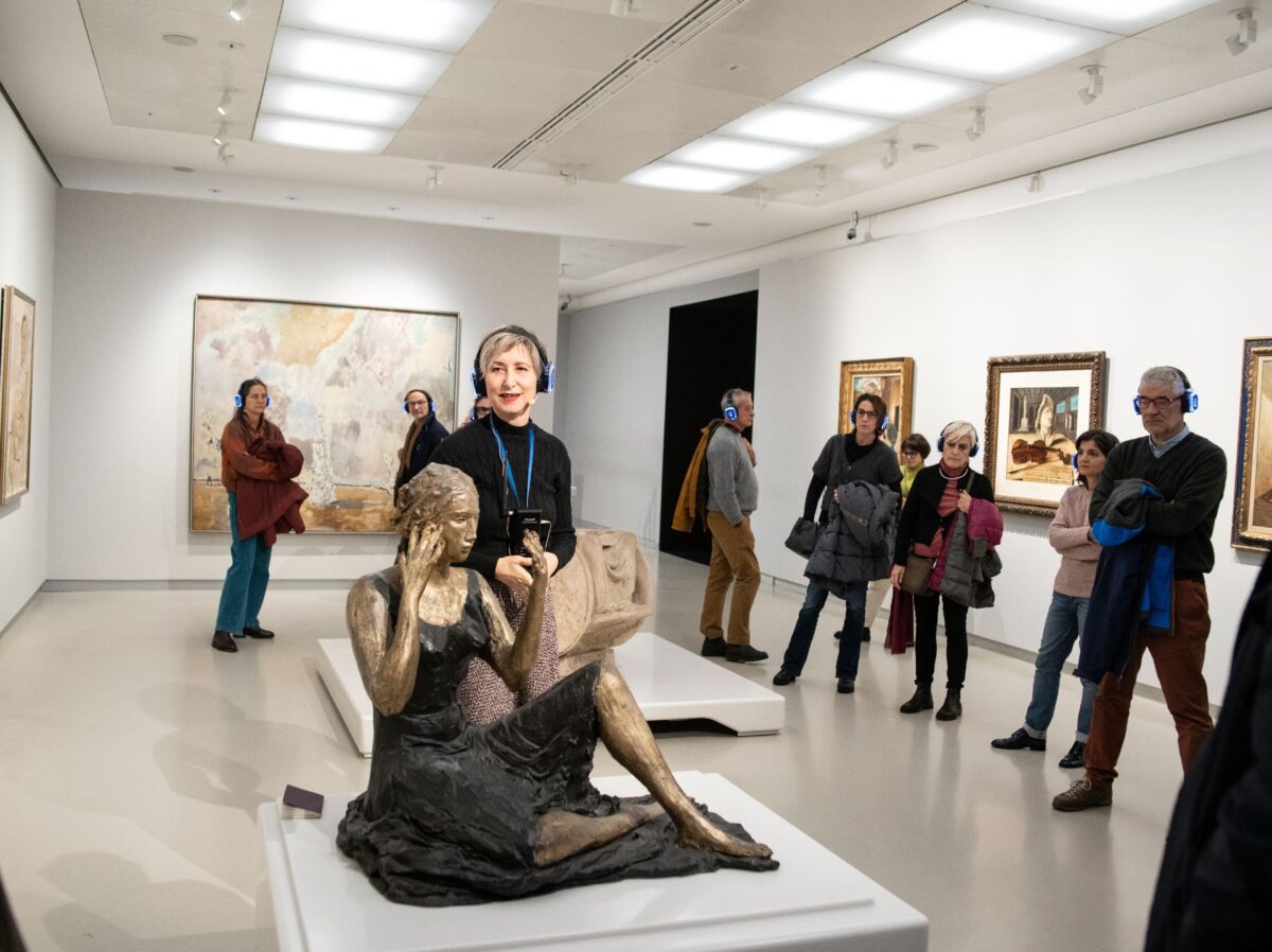 L'attrice e guida nelle sale del Museo del Novecento parla di fronte a un opera che raffigura una donna seduta con la pelle oro, il vestito nero nell'atto di specchiarsi. intorno all'attrice, il pubblico munito di cuffie