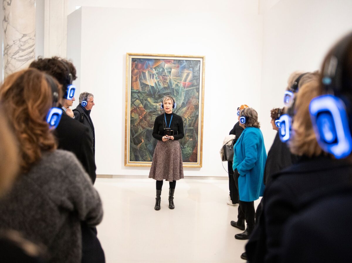 L'attrice sta con la cuffia di fronte a un quadro futurista di boccioni al museo del novecento, intorno a lei il pubblico in cuffia, in ascolto