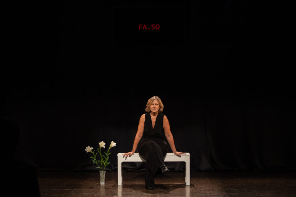 Una donna vestita di nero sta seduta su una panchina bianca, per terra accanto a lei un mazzo di gigli in un vaso di vetro. dietro proiettata la scritta falso, parte del gioco performativo dello spettacolo maternità