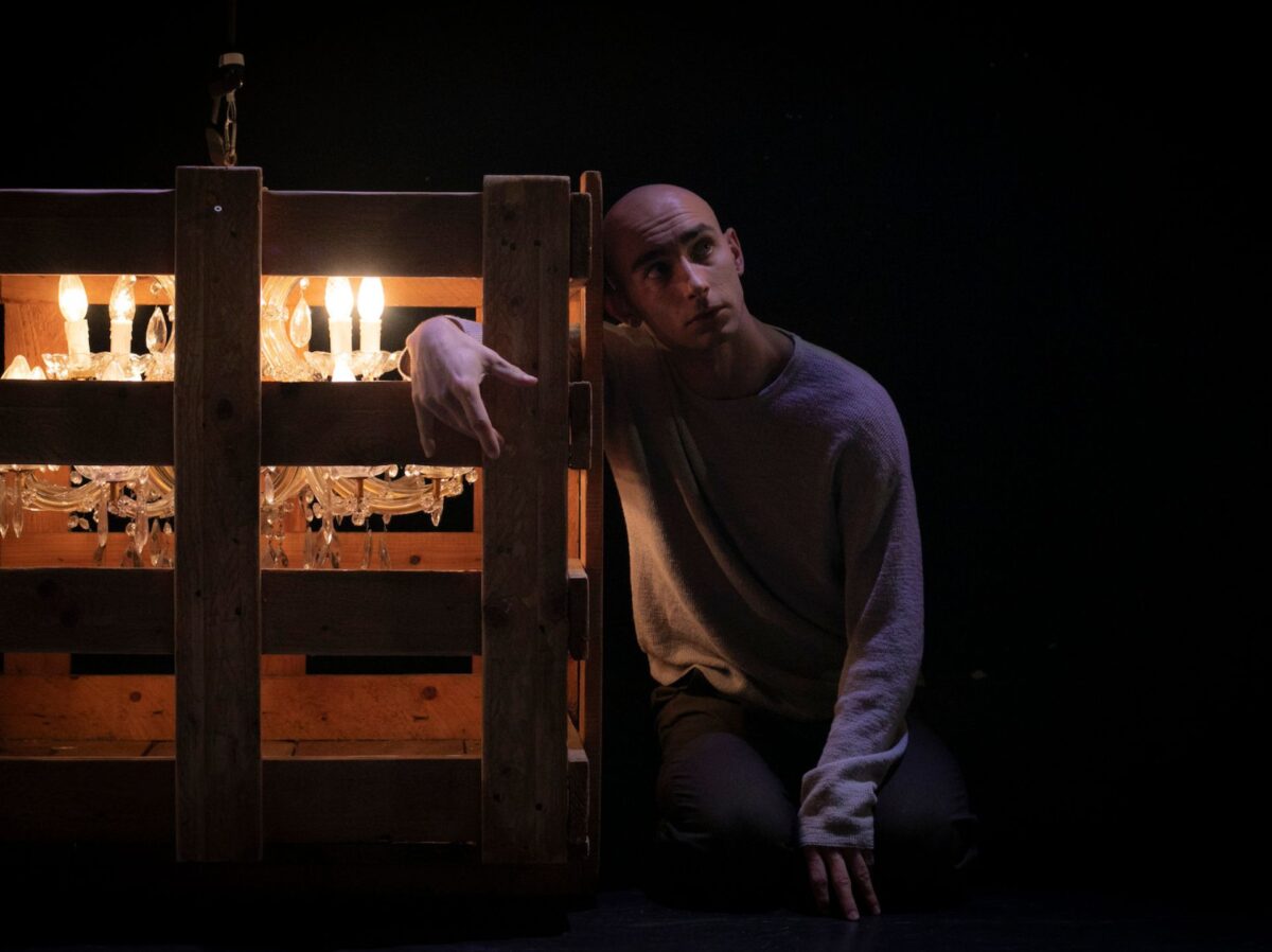 Un giovane sta sconsolato e pensieroso accanto a una struttura di pallet di legno, che contiene un lampadario a goccia che illumina la scena
