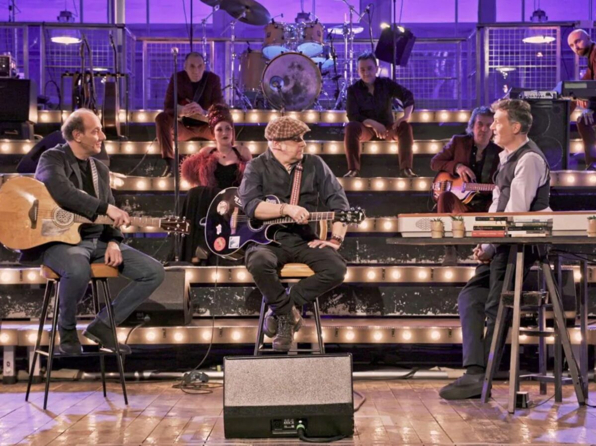In primo piano tre persone, che chiacchierano, due con la chitarra e un terzo appoggiato a una pianola, dietro altri musicisti su una gradinata con i gradini illuminati dai segnapassi, sul retro una grande vetrata illuminata da una luce violacea