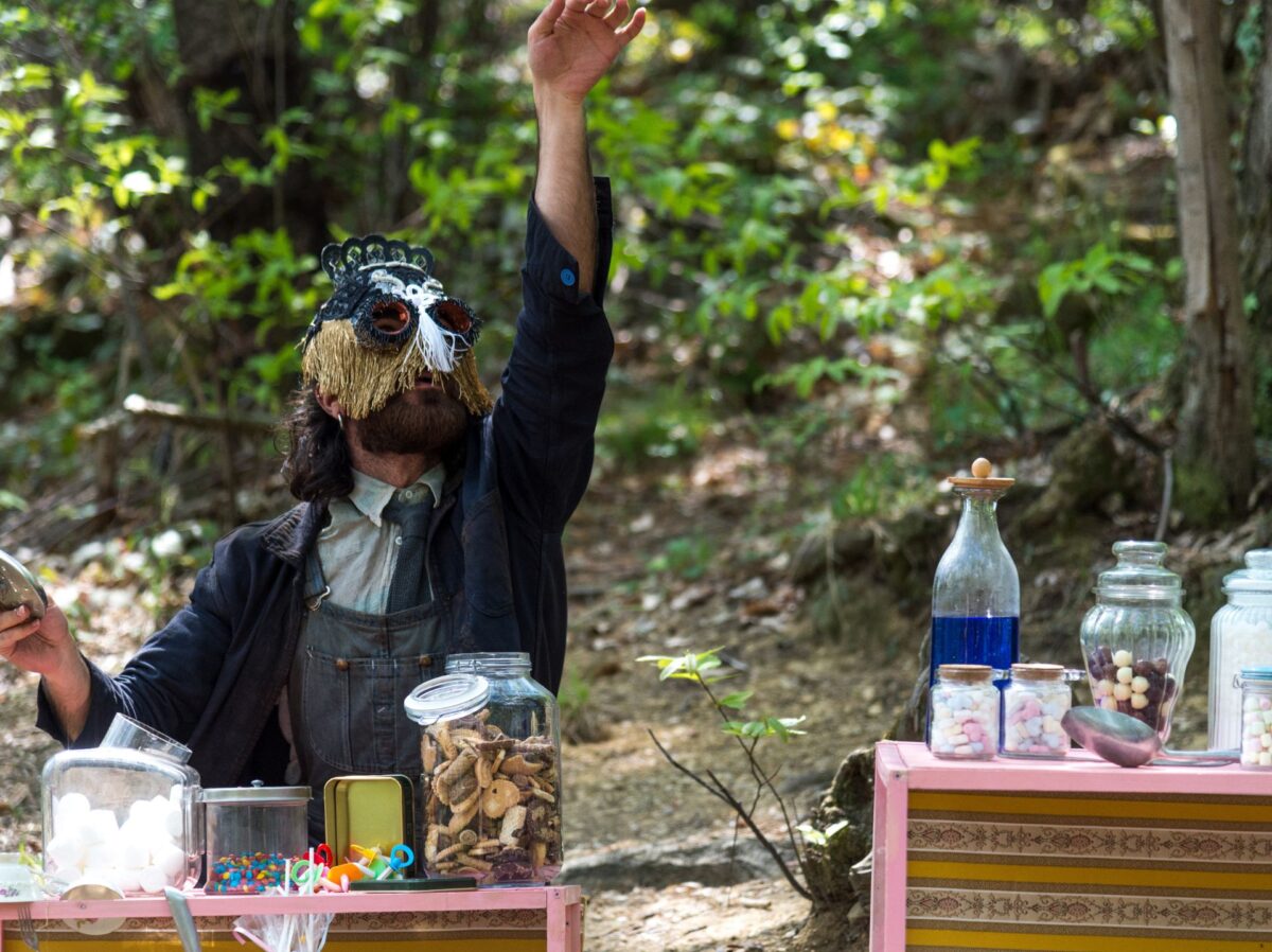 Una figura con maschera di pizzo in un bosco sta di fronte a tavoli pieni di recipienti con sassolini, caramelle, bottiglie con intrugli colorati, quasi fosse un alchimista