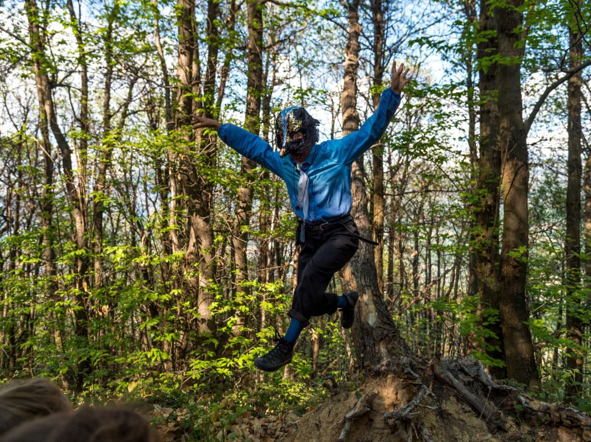 Una figura mascherata con camicia azzurra e pantaloni neri salta agilmente in un bosco con braccia e gambe spalancate