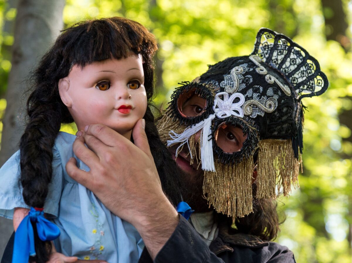 Una figura con una maschera di pizzo tiene sotto il mento con le mani una bambola dal vestitino azzurro