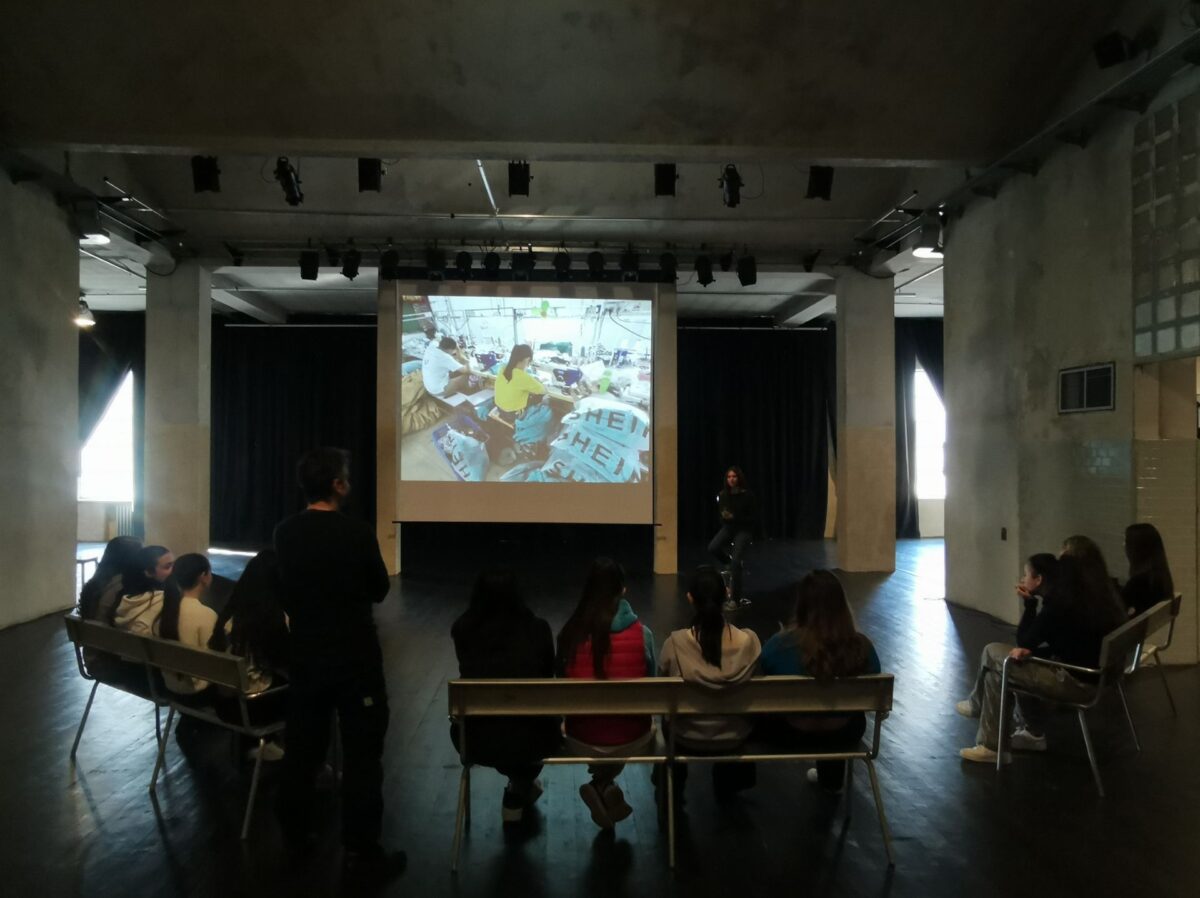 I ragazzi sono disposti su panche al centro della scena, sullo schermo proiettata l'immagine di lavoratrici del marchio fast fashion shein, nella struttura industriale dell'ex mensa dell'ex o.p. paolo pini, ora teatro