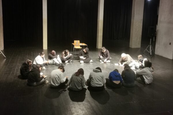 Un gruppo di adolescenti sta seduto in cerchio a teatro, chino sui quaderni a scrivere, in un incontro del laboratorio di scrittura poetica condotto da gigi gherzi