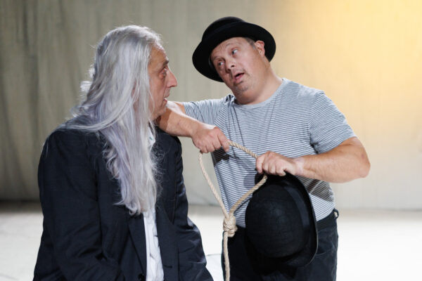 Un uomo dai capelli lunghi color argento guarda sbigottito un altro uomo con bombetta nera e tshirt grigia che tiene in mano un cappio fatto di corda, nella rappresentazione di due personaggi beckettiani nello spettacolo la buca