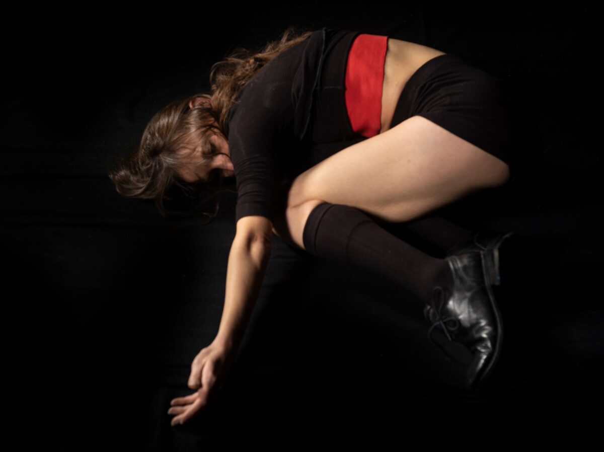 La danzatrice, con maglia nera e bordo rosso, mutande nere e un gambale nero, sta in posizione fetale su un pavimento nero, attorcigliata su se se stessa, quasi volesse rimpicciolirsi