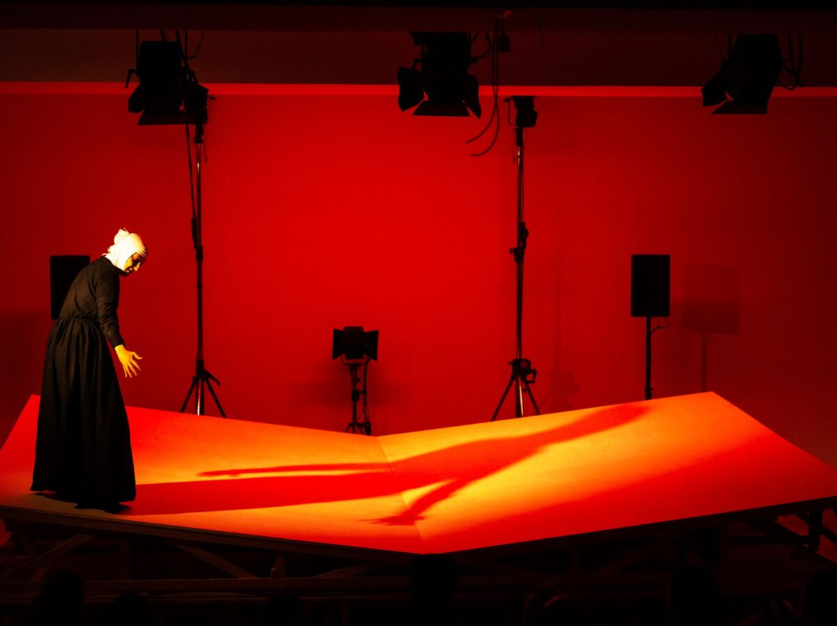 Un'attrice vestita da suora sta nell'angolo sinistro della foto, proiettando al sua ombra in una luce diffusa rossa, su una struttura a terra che riproduce un grosso libro aperto con due pagine bianche