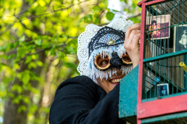 Una figura di donna con maschera di pizzo sta accanto ad una gabbia colorata di rosa e azzurro, con espressione sorniona e quasi malefica