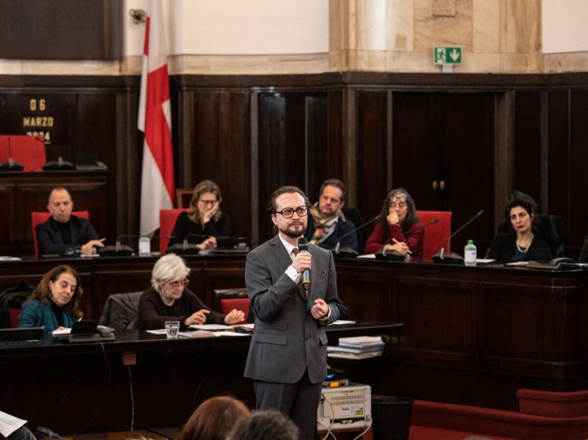 In primo piano, al centro dell'aula del consiglio comunale di palazzo marino, l'attore che interpreta primo levi, sul fondo sugli scranni il pubblico, in una disposizione semicircolare