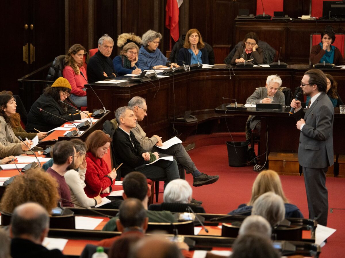 A lato dell'aula del consiglio comunale di palazzo marino, l'attore che interpreta primo levi, di fronte sugli scranni il pubblico, in una disposizione semicircolare