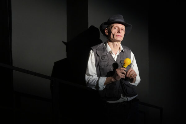 un uomo con un cappello, camicia e gilet mostra in primo piano una torcia di colore giallo, alle sue spalle la sua ombra, è l'evocazione dell'artista joseph beuys