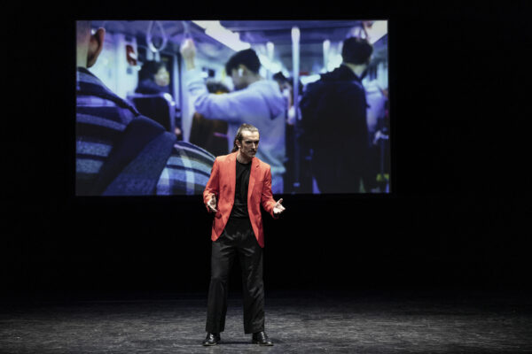 L'attore con giacca rossa sta al centro della scena, parlando e gesticolando, dietro di lui la proiezione di un momento quotidiana nel vagone della metropolitana